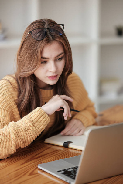 Eine schöne kaukasische Studentin studiert in der Ferne am College. Sie sitzt zu Hause mit Laptop und Notizblock an einem Tisch und schaut konzentriert einer Videokonferenzstunde zu. Hohe Qualität - Foto, Bild