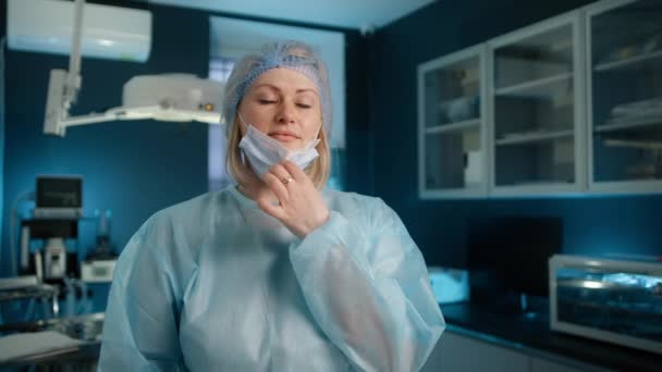 Confident female nurse in medical cabinet looking at camera and taking off face mask during pandemic. Portrait of smiling caucasian female doctor surgeon standing in hospital wearing protective mask - Footage, Video
