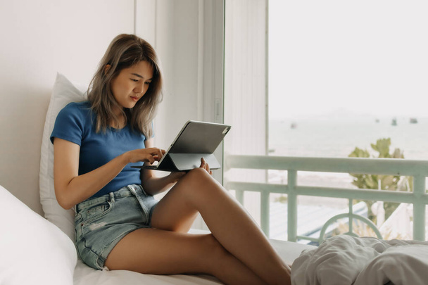 Mujer asiática seria trabajando en línea con la tableta en la cama del hotel en viaje - Foto, Imagen