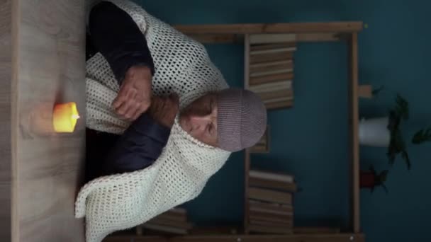 Oude man gekleed in een warme winterjas zit thuis aan een tafel en verwarmt zijn handen van een brandende kaars. Concept van geen verwarming in de winter thuis. Verticale video - Video