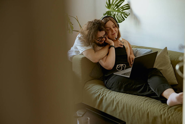 Mujer con auriculares trabajando en el portátil mientras su novio la abraza en casa. Concepto de trabajo a distancia - Foto, Imagen