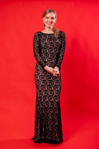 A beautiful young woman in a black dress poses standing isolated on a red background. - Valokuva, kuva