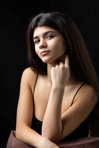 Classic studio portrait of a young brunette dressed in a black top, who is sitting on a chair against a black background. - Foto, Imagen