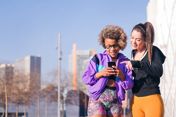 two sportswomen using a mobile phone before running standing on the city street, concept of friendship and sportive lifestyle - Foto, Bild