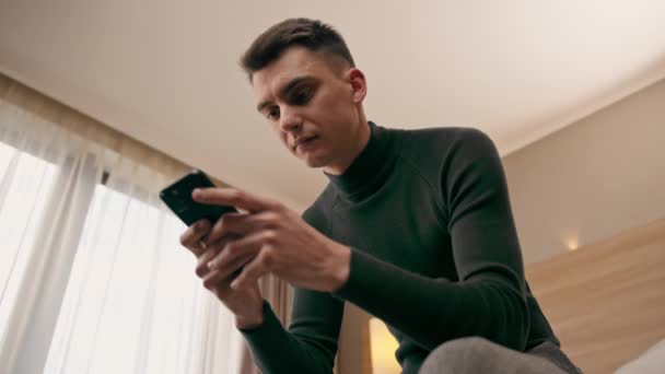 un joven en la habitación de una habitación de hotel de lujo tiene un teléfono inteligente en sus manos y resuelve problemas de negocios después del viaje - Metraje, vídeo