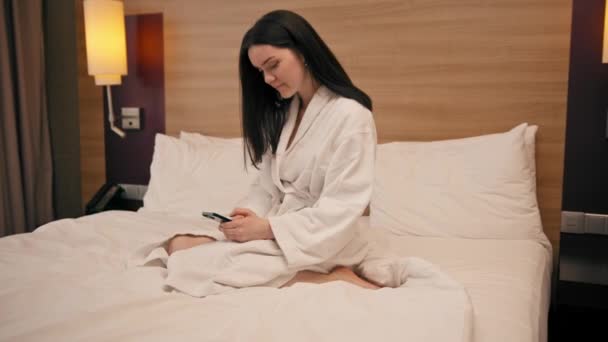mladá dívka v bílém plášti v luxusním hotelovém pokoji drží smartphone a řeší obchodní problémy po cestě - Záběry, video