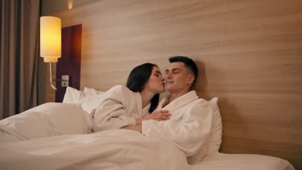 ハッピー恋人の旅行コンセプトにキスベッドの白いローブに横たわるホテルの部屋のハネムーンの肖像画若いカップル - 映像、動画