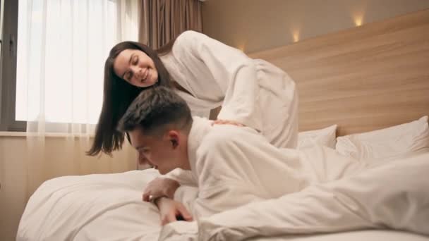 портрет молодої пари на медовий місяць в готельному номері лежить ліжко в білих халатах флірт сміється щасливі коханці - Кадри, відео
