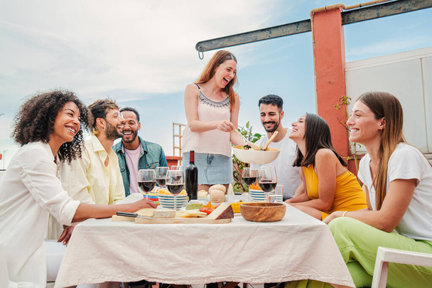 Caucasian joyful woman gathered with her friends, serving a lunch sitting at table talking and drinking wine ,enjoying a great time together. Group of young adult people celebrating at home rooftop - Фото, изображение