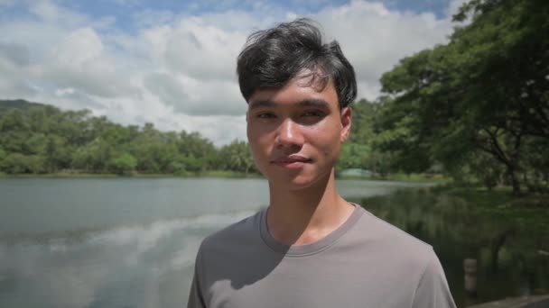 Retrato asiático adolescente guapo niño sonriendo y mirando a la cámara mientras está de pie en el lago natural en el parque público. Joven estilo de vida adolescente. Búsqueda al aire libre. - Metraje, vídeo