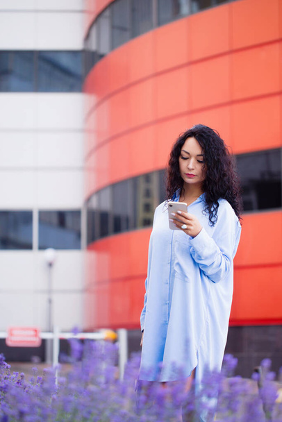 Портрет красивой офисной работницы в длинной синей рубашке со смартфоном на фоне городского пейзажа - Фото, изображение