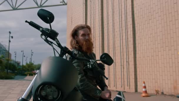 ひげを持つ残忍な赤毛のバイカーは,レースの開始前に彼の手の渇きのスピードでヘルメットを保持する黒いオートバイに座っています - 映像、動画