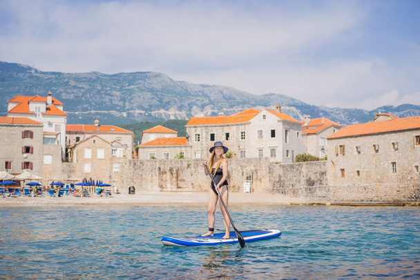 Mujeres jóvenes se divierten de pie remando en el mar de agua azul en Montenegro. En el contexto del casco antiguo de Budva. SUP. - Foto, Imagen
