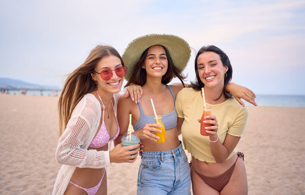 Happy portrait of three young Caucasian women on beach together hugging looking at camera cheerful. Smiling girls friends holding fruit smoothies enjoying their summer vacation in Mediterranean. - Fotó, kép