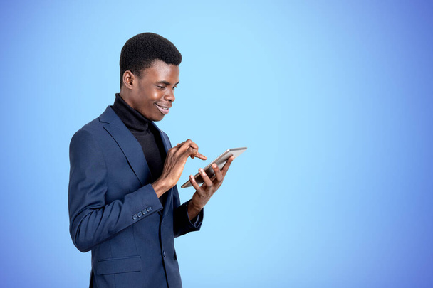 Handsome African American businessman wearing formal suit is standing watching at tablet device near empty blue wall in background. Concept of modern gadgets, mobile communication, internet exploring - Foto, Imagen
