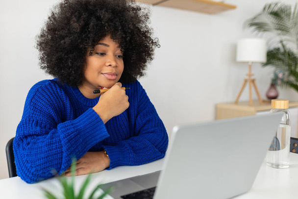 Молодая африканская американка, работающая над ноутбуком. Сконцентрированная деловая женщина смотрит на экран компьютера, читая какую-то статью о работе или слушая на виртуальных уроках видеозвонков. Концепция бизнеса - Фото, изображение