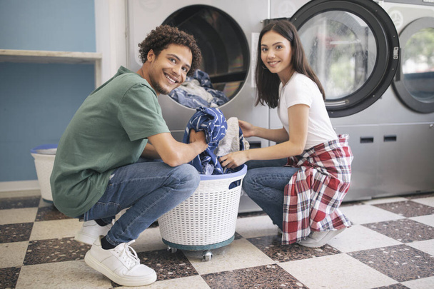 Happy Young Multicultural Couple Doing Laundry Together Loading Washer Machine At Laundromat Room. Guy Washing His Clothes And Posing Smiling At Camera. Public Laundrette Service - Foto, imagen