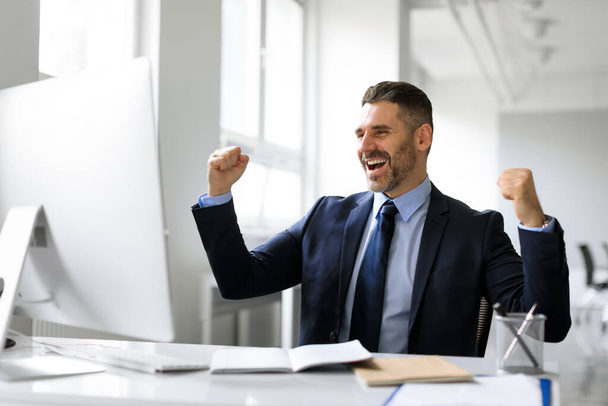 オフィスのコンピュータの前で成功を祝う興奮した中年のビジネスマン,男性起業家は拳を振るとはい叫んで,良いニュースに反応します。 - 写真・画像