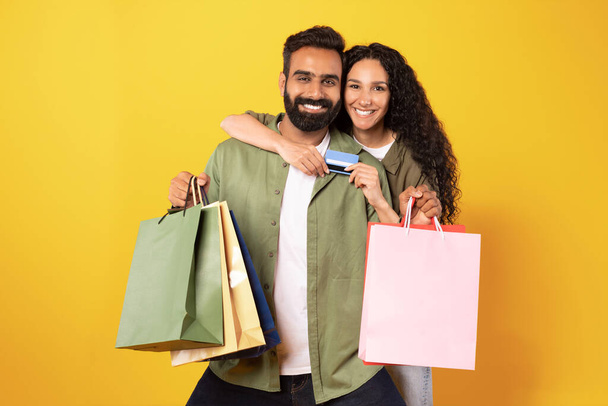 Nákupní nabídka. Portrét Happy Arabic Zákazníci Pár s kreditní kartou a nákupní tašky, Objímání a reklama Sezónní prodej přes žluté pozadí. Studio záběr spokojených kupujících - Fotografie, Obrázek