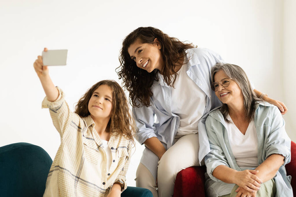 Glad european adult, senior women and teen girl sit on sofa, make selfie on smartphone in living room interior. Family, photo female generation, relationship mom, grandma and daughter, video call - Фото, изображение