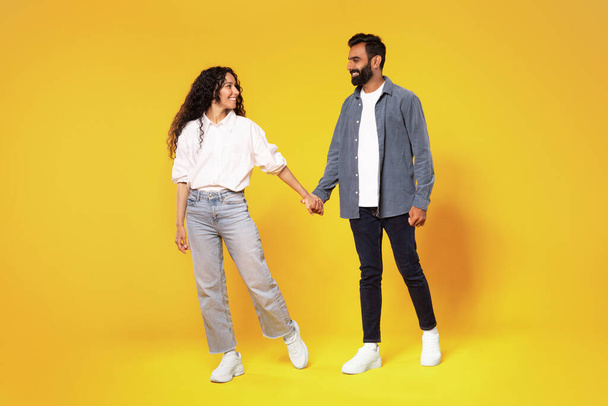 Romantische Beziehung. Glückliches arabisches Millennial-Paar, das sich Händchen haltend über gelbem Studiohintergrund in voller Länge anlächelt. Eheleute Mann und Frau posieren gemeinsam. Familie, Liebe - Foto, Bild