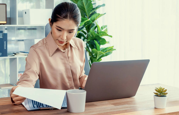 Młoda azjatycka entuzjastyczna bizneswoman w nowoczesnym biurze przy użyciu laptopa do pracy i pisania notatek. Staranna i atrakcyjna kobieta biurowa pracująca nad notebookiem komputerowym w swojej przestrzeni biurowej. - Zdjęcie, obraz