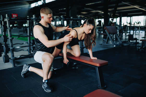 In the gym, a skilled male personal trainer aids a woman in her weightlifting journey, specifically focusing on her dumbbell lift and technique. - Фото, зображення