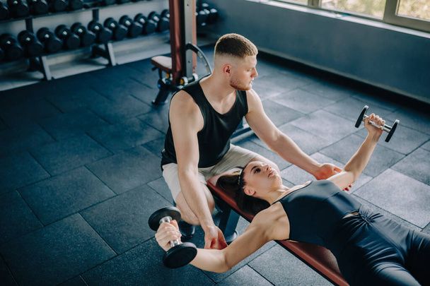 Az edzőteremben egy hozzáértő férfi személyi edző útmutatást nyújt egy súlyzót emelő nőnek. Fitness képzés Férfi személyi edző segít a nőnek a súlyzós testmozgás - Fotó, kép
