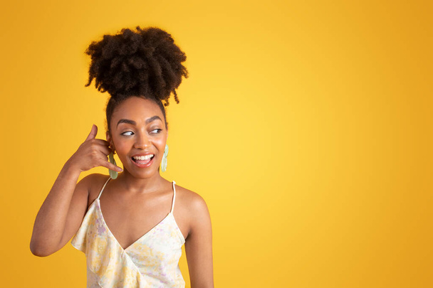 Cheerful millennial african american lady in dress making call me gesture, flirting, isolated on yellow background, studio. Good news, gossip, communication sign, ad and offer - Foto, Imagen