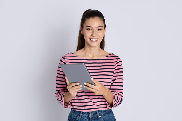 Happy pretty brunette long-haired millennil woman wearing casual outfit using modern pc digital tablet on grey studio background, checking newest app, working online, copy space - Photo, image