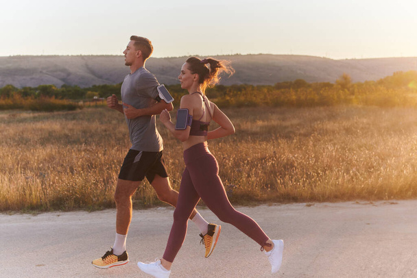 A handsome young couple running together during the early morning hours, with the mesmerizing sunrise casting a warm glow, symbolizing their shared love and vitality.  - Zdjęcie, obraz