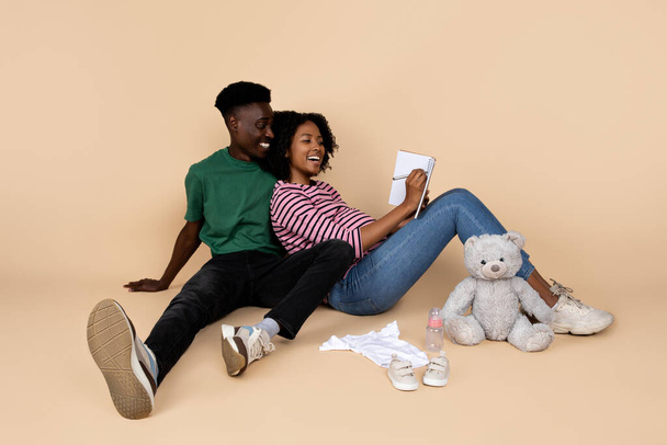 Positive young african american guy and woman with big belly sit on floor with baby things and toys, make notes in diary, isolated on beige background. Planning, relationships, expect of child - Photo, Image