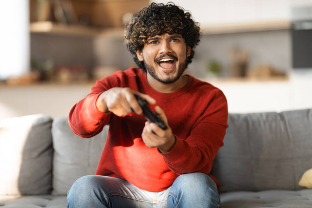 Concepto de competencia en línea. Retrato del joven indio emocionado jugando videojuegos en casa, alegre consola masculina oriental sosteniendo el joystick, jugador sentado en el sofá en la sala de estar, espacio para copiar - Foto, Imagen