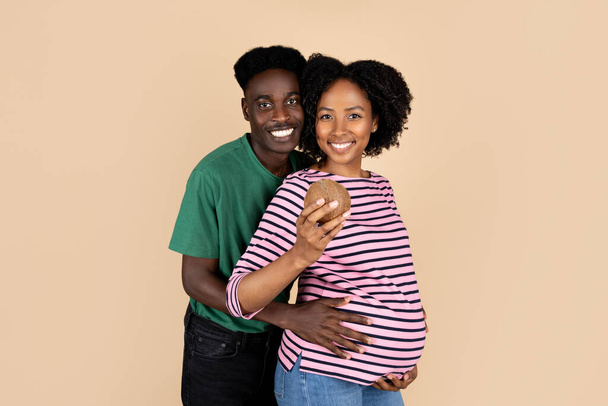 Sorridente marito afro-americano millenario che abbraccia una donna con la pancia grande, signora che tiene la noce di cocco, isolata su sfondo beige, studio. Assistenza sanitaria, famiglia, attesa bambino, annuncio e offerta - Foto, immagini