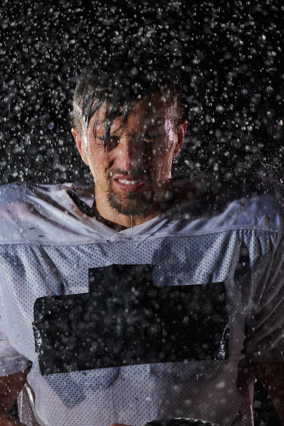 American Football Field: Samotny wojownik atleta stojący na polu trzyma swój kask i gotowy do gry. Gracz przygotowuje się do uruchomienia, ataku i przyziemienia Ocena. Deszczowa noc z dramatyczną mgłą, niebieskie światło. - Zdjęcie, obraz