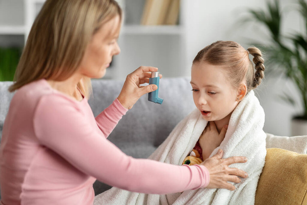 Preventing Attack. Caring mother giving blue asthma inhaler to her sick child at home, loving young mom taking care about ill little daughter suffering breathing problem, closeup shot - Photo, Image