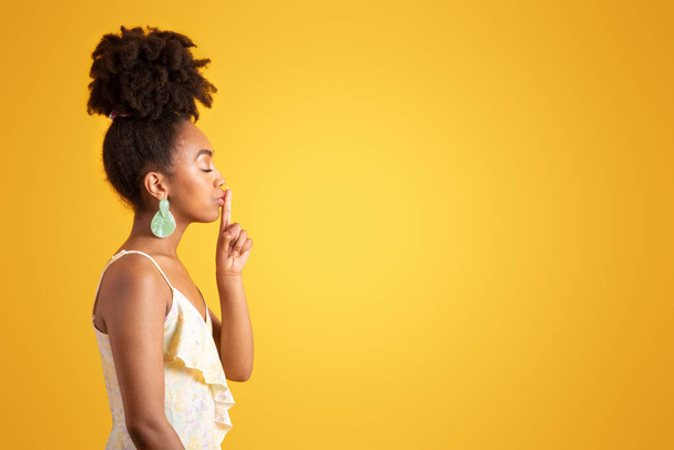 Lächelnde ruhige afrikanische Amerikanerin im Kleid, die eine shh-Geste macht, Finger an Lippen legt, isoliert auf gelbem Hintergrund, Studio, Profil. Lifestyle, Geheimnis, gute Nachrichten, Werbung und Angebot, Klatsch und Tratsch - Foto, Bild