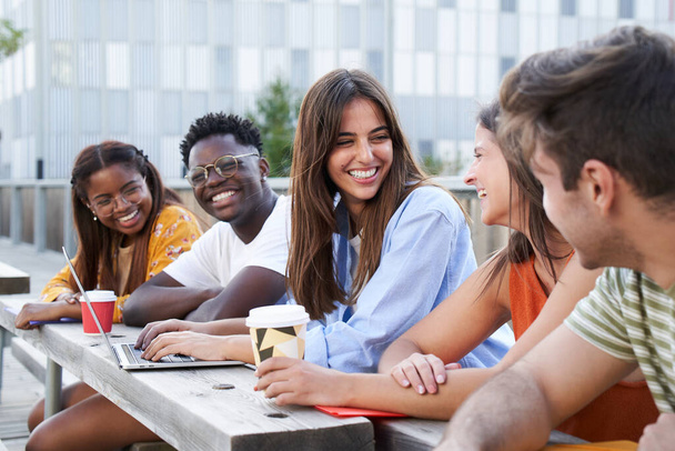 学生のグループは、大学のキャンパスで屋外に座って笑顔。1人の女の子はラップトップを使用しており、もう1人はコーヒーを飲む。それは多様な国の多人種集団です. - 写真・画像