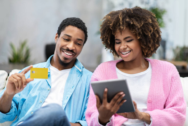 Веселые красивые африканские влюбленные американские юноши и девушки сидят дома на диване, используя цифровой планшет, держа в руках желтую пластиковую банковскую карточку. Интернет-банкинг - Фото, изображение