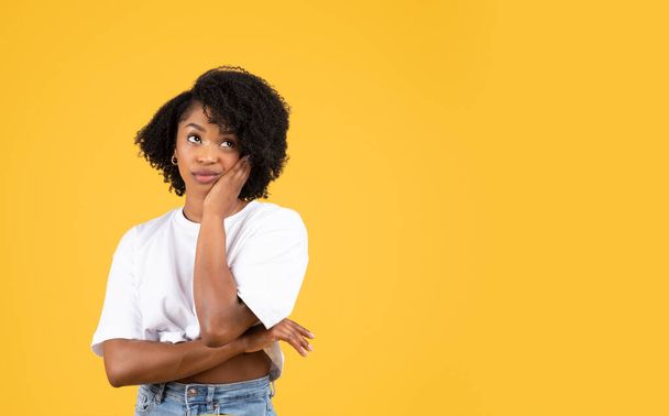 Sad pensive millennial black woman in casual looks at copy space, think, touch cheek, isolated on orange studio background. Choice, human emotion, ad and offer, teeth pain and health care - Photo, Image