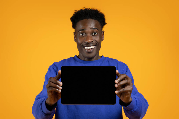 Wesoły przystojny młody czarny facet w luźnym stroju pokazując poziome nowoczesne PC cyfrowy tablet z pustym ekranem, pokazując ładne aplikacja, makieta, żółte tło studio - Zdjęcie, obraz