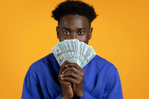 Портрет молодого афроамериканця в повсякденному синьому светрі з грошовими купюрами. Чорний хлопець отримав готівку, виграв гроші, ізольований на жовтому студійному фоні - Фото, зображення