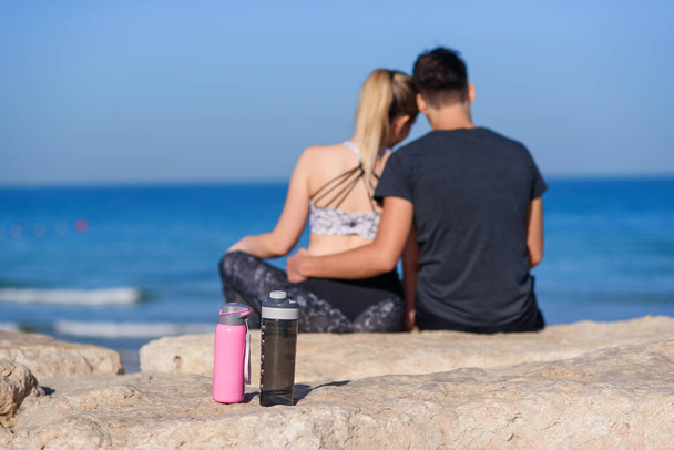 Молодая влюбленная пара сидит на скалистом пляже и смотрит на горизонт моря. Бутылки для воды на переднем плане. - Фото, изображение