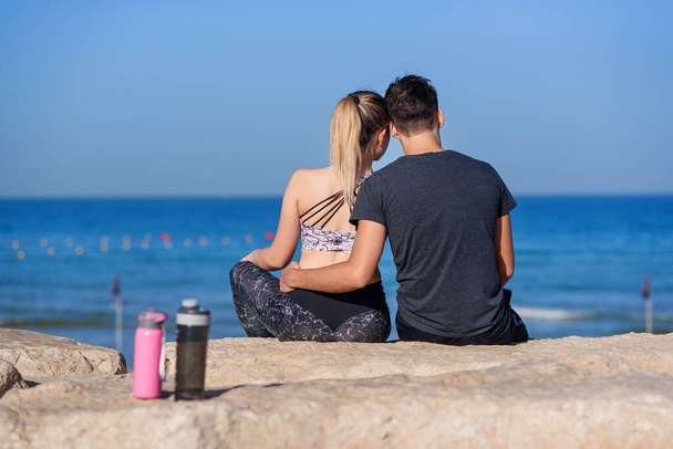 Ein junges verliebtes Paar sitzt am felsigen Strand und blickt am Horizont auf das Meer. Wasserflaschen im Vordergrund. - Foto, Bild