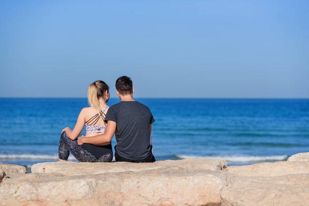 Ein junges verliebtes Paar sitzt am felsigen Strand und blickt am Horizont auf das Meer. Lovestory-Konzept. - Foto, Bild