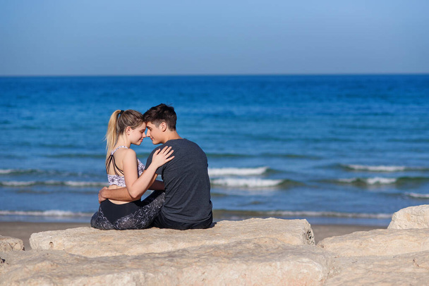 Young couple in love sitting on the rocky beach and looking on the horizon at sea. Lovestory concept. - Фото, изображение