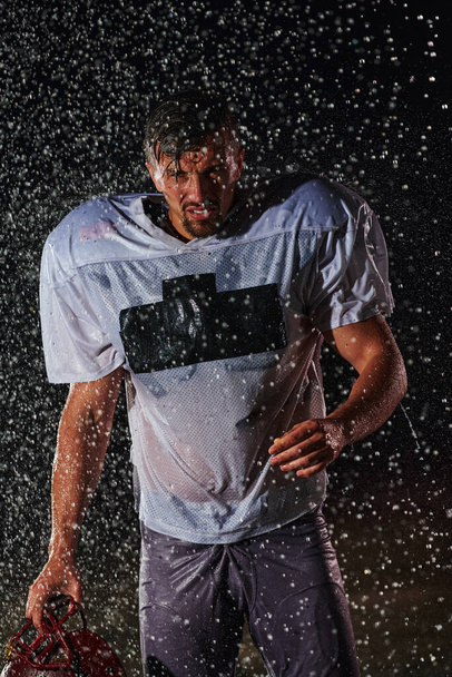 American Football Field: Lonely Athlete Warrior Standing on a Field Holds his Helmet and Ready to Play. Player Preparing to Run, Attack and Score Touchdown. Rainy Night with Dramatic Fog, Blue Light. - 写真・画像