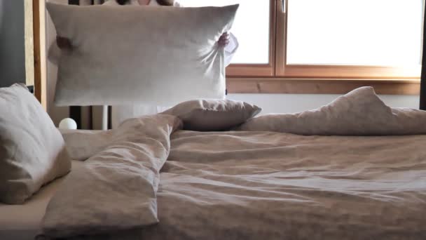 Bettschminken.Frauenhände richten die Kissen auf dem Bett neben dem Fenster auf. Frau im weißen Bademantel macht das Bett. Morgenreinigung im Schlafzimmer. 4k Filmmaterial - Filmmaterial, Video