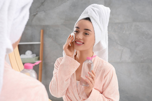 Beautiful woman in terry towel removing makeup with cotton pad near mirror indoors - Zdjęcie, obraz