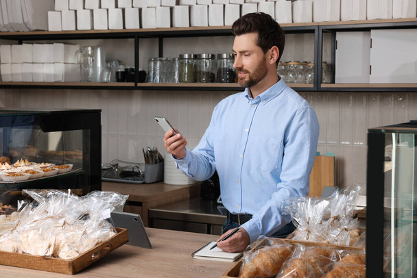 Handsome business owner with smartphone and pen at cashier desk in bakery shop - Photo, Image